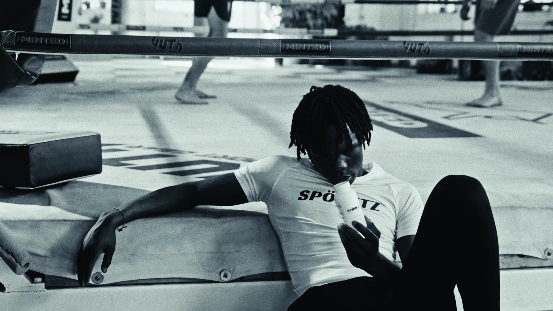 Grappler resting ringside in a white Spöretz tee, holding the MB40 bottle, black-and-white